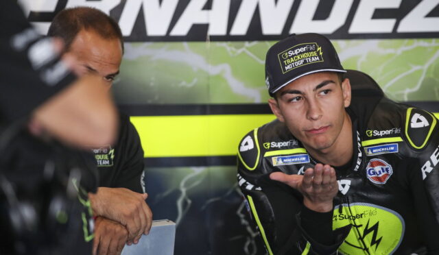 Motocross racer in neon-yellow and black gear sits in a team garage, palm raised as he talks to a teammate beside him.【informative】