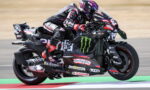 Motorcycle racer in pink helmet and black racing suit leaning into a high-speed corner on a Monster Energy-liveried bike with sponsor decals on the bodywork.