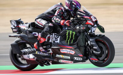 Motorcycle racer in pink helmet and black racing suit leaning into a high-speed corner on a Monster Energy-liveried bike with sponsor decals on the bodywork.