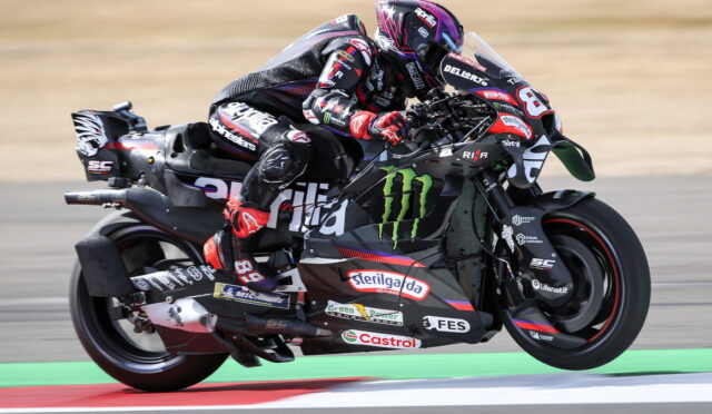 Motorcycle racer in pink helmet and black racing suit leaning into a high-speed corner on a Monster Energy-liveried bike with sponsor decals on the bodywork.