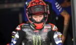 MotoGP rider in a red helmet and Yamaha Monster Energy racing suit preparing in the pit area, focused and ready to race, with sponsor logos visible.