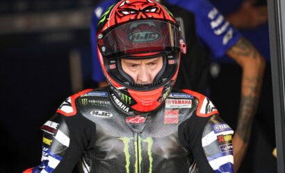 MotoGP rider in a red helmet and Yamaha Monster Energy racing suit preparing in the pit area, focused and ready to race, with sponsor logos visible.