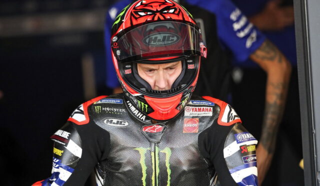 MotoGP rider in a red helmet and Yamaha Monster Energy racing suit preparing in the pit area, focused and ready to race, with sponsor logos visible.