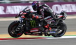 MotoGP rider on a black Aprilia sportbike speeding through a turn on a racetrack.