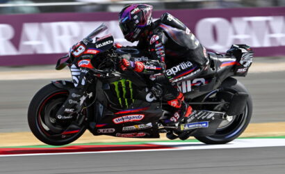 MotoGP rider on a black Aprilia sportbike speeding through a turn on a racetrack.
