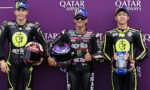 Three MotoGP racers in black neon-yellow Aprilia suits pose for a photo against a purple Qatar Airways backdrop, each holding a colorfully painted helmet.