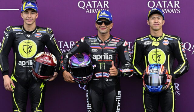 Three MotoGP racers in black neon-yellow Aprilia suits pose for a photo against a purple Qatar Airways backdrop, each holding a colorfully painted helmet.