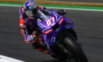 Motorcycle racer leaning into a sharp turn on a purple racing bike with sponsor decals on a track surface.