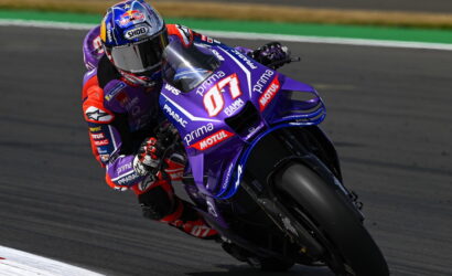 Motorcycle racer leaning into a sharp turn on a purple racing bike with sponsor decals on a track surface.