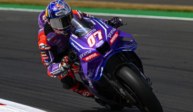Motorcycle racer leaning into a sharp turn on a purple racing bike with sponsor decals on a track surface.