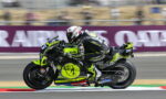 Motorcycle racer in neon yellow/black suit riding an Aprilia bike at a race track, speed blur behind the rider.