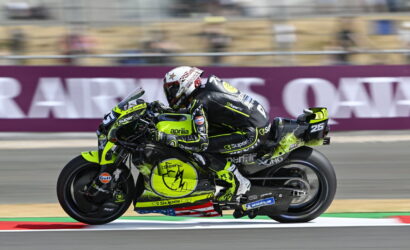 Motorcycle racer in neon yellow/black suit riding an Aprilia bike at a race track, speed blur behind the rider.