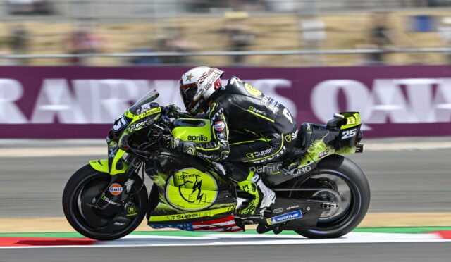 Motorcycle racer in neon yellow/black suit riding an Aprilia bike at a race track, speed blur behind the rider.