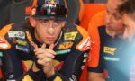 MotoGP rider in KTM leathers with a nose bandage sits with hands clasped, flanked by teammates in orange in a paddock.