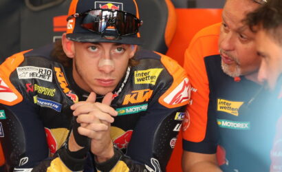 MotoGP rider in KTM leathers with a nose bandage sits with hands clasped, flanked by teammates in orange in a paddock.