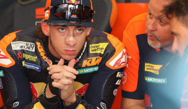 MotoGP rider in KTM leathers with a nose bandage sits with hands clasped, flanked by teammates in orange in a paddock.