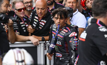 MotoGP rider in a black racing suit drinks from a blue bottle as photographers crowd around behind a barrier.