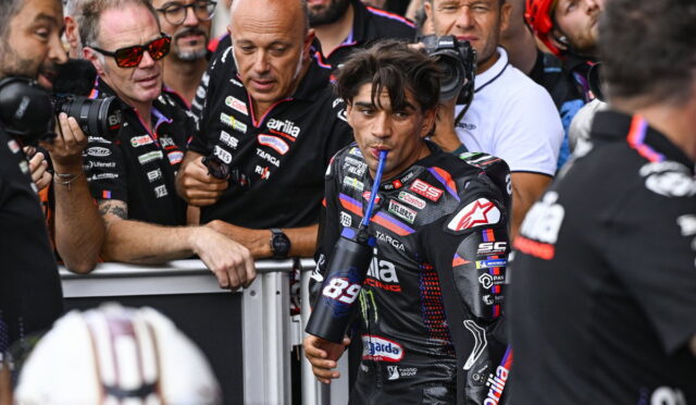 MotoGP rider in a black racing suit drinks from a blue bottle as photographers crowd around behind a barrier.