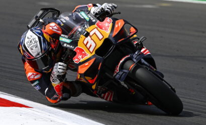 MotoGP rider in orange gear leans into a left turn on a race track; bike #37 with Red Bull branding.