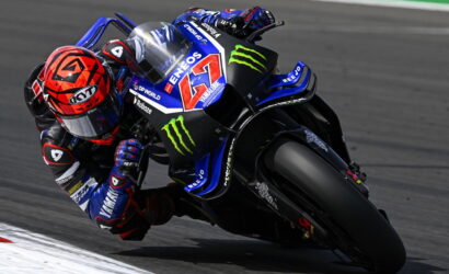 Racing motorcycle leaning into a left-hand turn, rider in blue and black suit on a Yamaha bike with Monster Energy logos and the number 46 visible on the fairing, track surface blurred in the background.