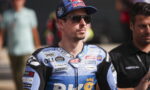 Man wearing a blue Red Bull cap and dark sunglasses, in a blue racing suit covered with sponsor logos, likely at a race event in a paddock area.