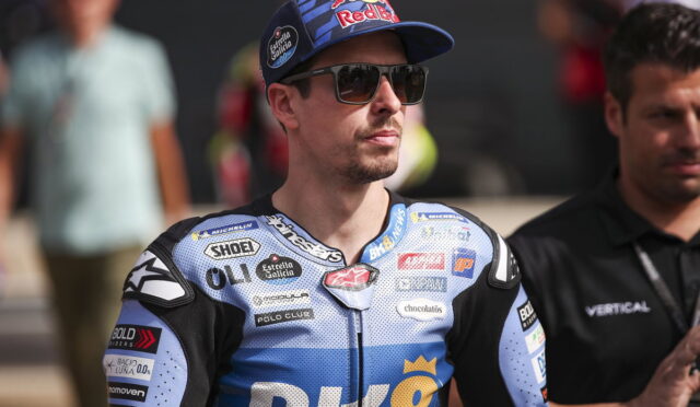 Man wearing a blue Red Bull cap and dark sunglasses, in a blue racing suit covered with sponsor logos, likely at a race event in a paddock area.