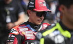 MotoGP rider in a red racing suit and cap with sponsor patches, wearing dark sunglasses in the paddock.