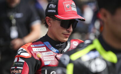 MotoGP rider in a red racing suit and cap with sponsor patches, wearing dark sunglasses in the paddock.