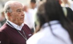 Older man with a maroon sweater and lanyard listening to someone in a crowded outdoor event.