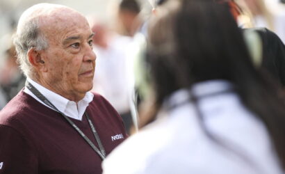 Older man with a maroon sweater and lanyard listening to someone in a crowded outdoor event.