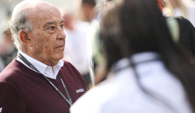 Older man with a maroon sweater and lanyard listening to someone in a crowded outdoor event.