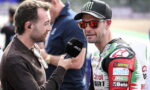 Reporter interviews a motorcyclist in racing suit with sponsor logos at a track, microphone close to his mouth.
