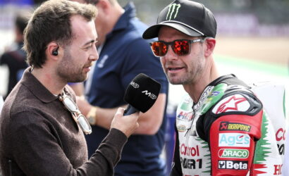 Reporter interviews a motorcyclist in racing suit with sponsor logos at a track, microphone close to his mouth.