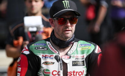 Motorcycle racer in green-white-red riding suit and black Monster cap, red sunglasses, standing in pit area with crew in background (Castrol logos visible).