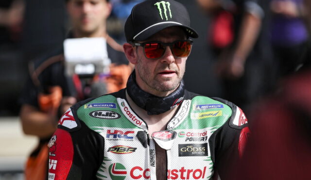 Motorcycle racer in green-white-red riding suit and black Monster cap, red sunglasses, standing in pit area with crew in background (Castrol logos visible).