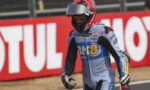 Motocross rider in blue race suit and red helmet walks along track with red advertising banner behind.