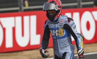 Motocross rider in blue race suit and red helmet walks along track with red advertising banner behind.