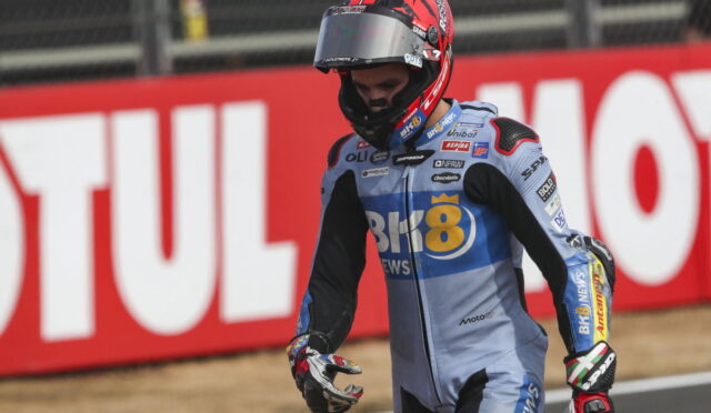 Motocross rider in blue race suit and red helmet walks along track with red advertising banner behind.