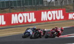 Motorcycle racers lean into a sharp turn on a racetrack, red Motul banners lining the fence in the background.