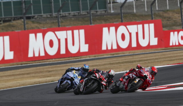 Motorcycle racers lean into a sharp turn on a racetrack, red Motul banners lining the fence in the background.