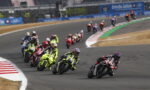 Group of motorcycle racers lean into a curve on a race track, with a black-red rider leading the pack.
