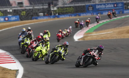 Group of motorcycle racers lean into a curve on a race track, with a black-red rider leading the pack.