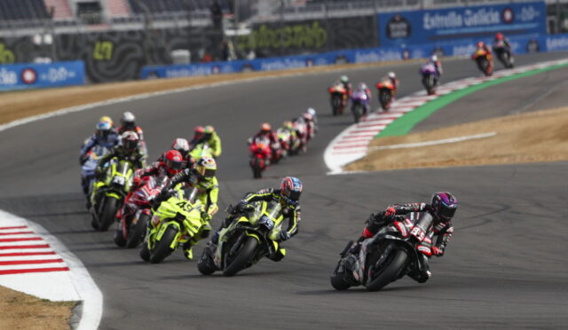 Group of motorcycle racers lean into a curve on a race track, with a black-red rider leading the pack.