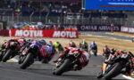 Group of motorcycle racers leaning into a sharp turn on a race track, dust kicked up behind them.