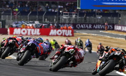 Group of motorcycle racers leaning into a sharp turn on a race track, dust kicked up behind them.