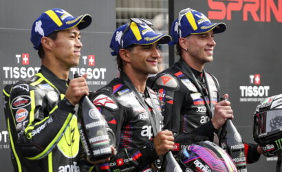 Three motorcycle racers stand on a podium holding champagne bottles, wearing racing suits and blue-yellow caps with sponsor logos.
