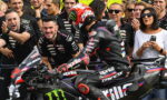 Motorcycle racer in black Aprilia leathers on a black race bike with Monster energy branding, surrounded by cheering pit crew and spectators behind a barrier.
