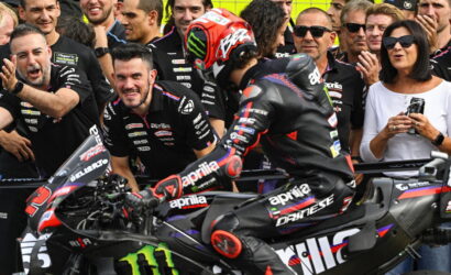 Motorcycle racer in black Aprilia leathers on a black race bike with Monster energy branding, surrounded by cheering pit crew and spectators behind a barrier.