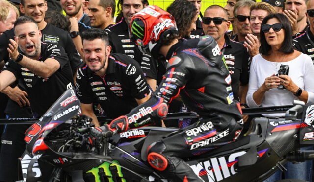 Motorcycle racer in black Aprilia leathers on a black race bike with Monster energy branding, surrounded by cheering pit crew and spectators behind a barrier.