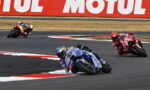 Motorcycle race taking a sharp left turn; blue bike leads, red bikes behind on a curving track with a large MOTUL banner in the background.